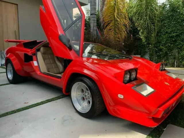 Lamborghini Countach 5000 S Prova Replica - Engine Stories Red sports car with iconic scissor doors open, parked in a driveway surrounded by lush greenery.