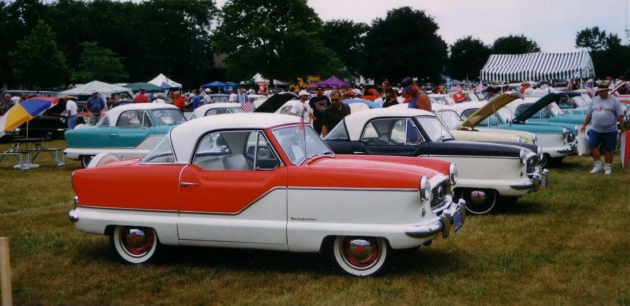 Vintage car show featuring classic red and white Nash Metropolitan models on display, surrounded by enthusiasts and vendors.