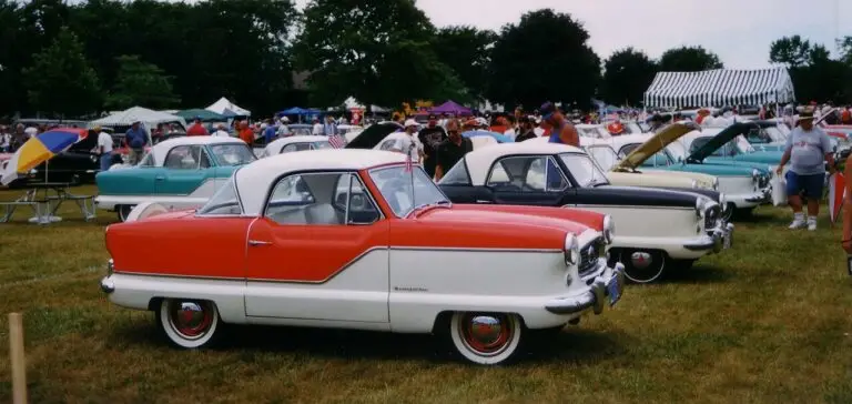 Vintage car show featuring classic red and white Nash Metropolitan models on display, surrounded by enthusiasts and vendors.