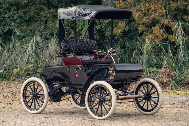Oldsmobile Curved Dash - 1901 - Engine Stories Vintage open-top car with black canopy, thin white tires, and brass details, parked outdoors on a leafy path.