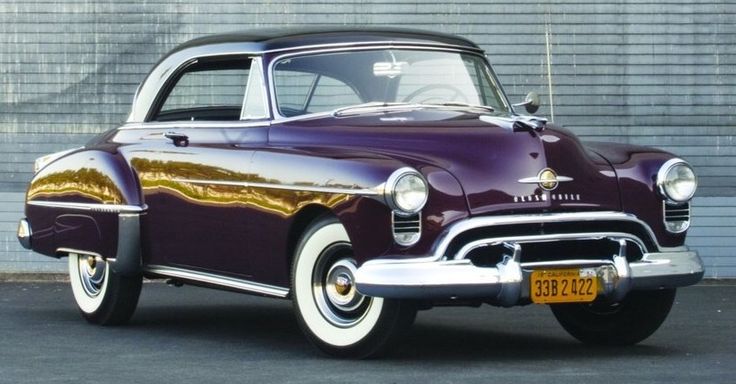 Oldsmobile Rocket 88 1950 - Engine Stories Vintage purple classic car with chrome details and whitewall tires parked in front of a textured wall.