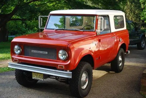 Red vintage off-road vehicle parked outdoors on a sunny day, with trees in the background. Classic SUV with white roof.