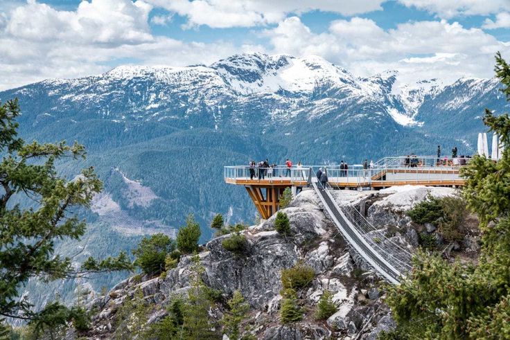 Scenic view of a mountain observation deck with snow-capped peaks in the background under a cloudy sky, surrounded by lush greenery.