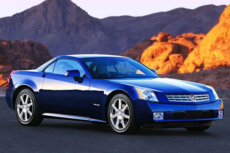 The Cadillac XLR was no Luxury Corvette - Engine Stories Sleek blue sports car parked against a scenic desert mountain backdrop at sunset.