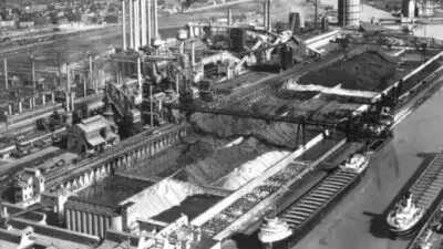 Aerial view of a bustling industrial steel plant with smokestacks, large storage areas, and docked cargo ships along a river canal.