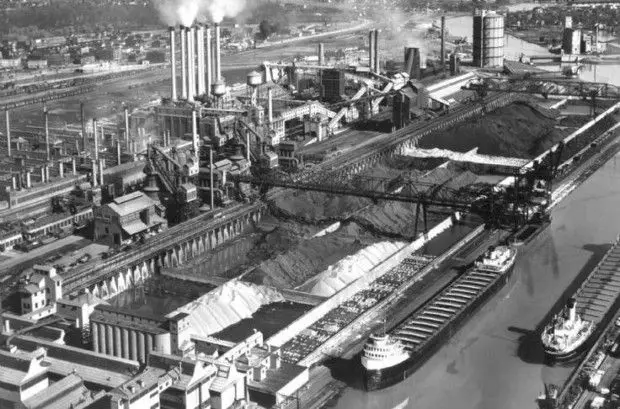 Aerial view of a bustling industrial steel plant with smokestacks, large storage areas, and docked cargo ships along a river canal.