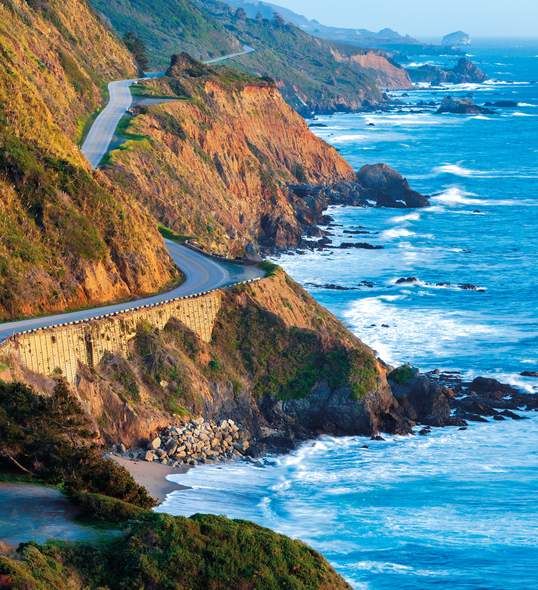 Scenic coastal road along rugged cliffs and ocean waves on a sunny day, showcasing the natural beauty of a picturesque landscape.