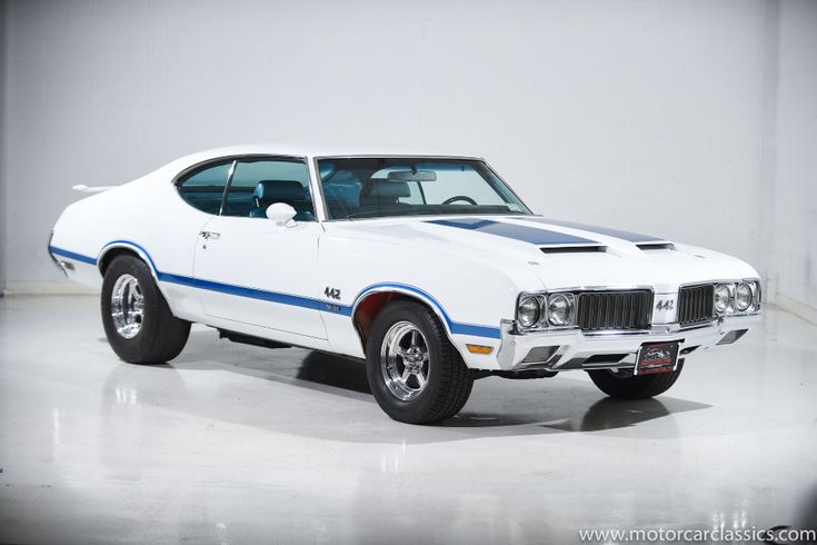 White classic Oldsmobile 442 with blue racing stripes in a showroom setting, showcasing vintage automotive design.