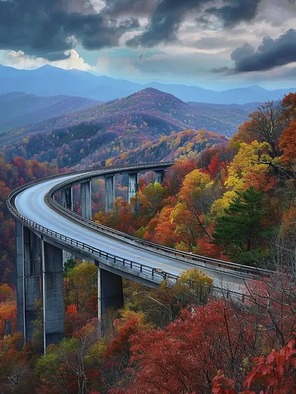 Curving mountain highway through vibrant autumn foliage under a dramatic sky, offering a scenic view of nature's beauty.