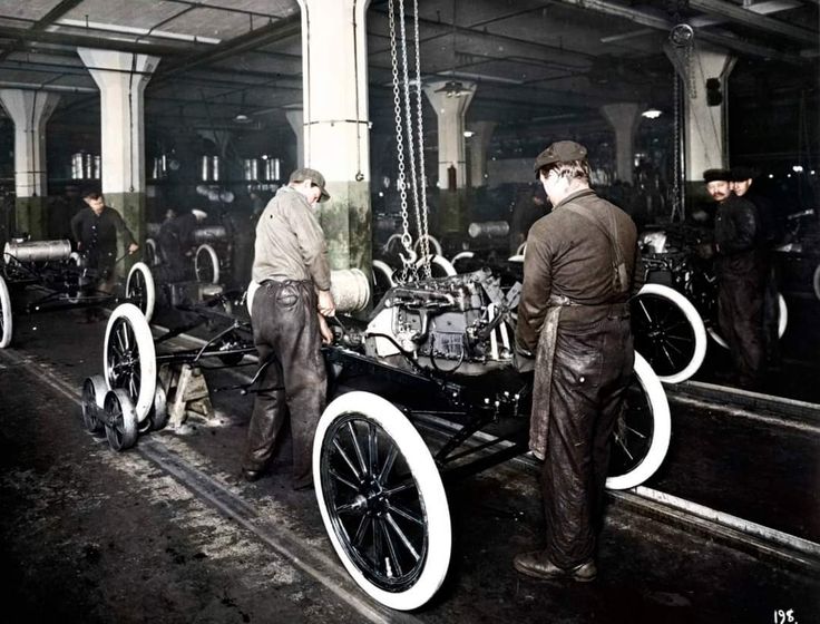 Workers assembling vintage car chassis in an early 20th-century factory setting.