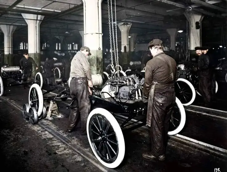 Workers assembling a Model T Ford at the Highland - Engine Stories Workers assembling vintage car chassis in an early 20th-century factory setting.