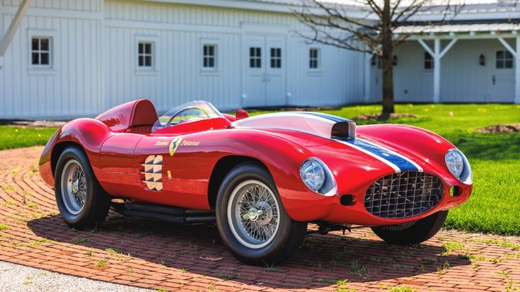 Red vintage racing car parked on a brick driveway in front of a white barn, showcasing classic design and vibrant color.