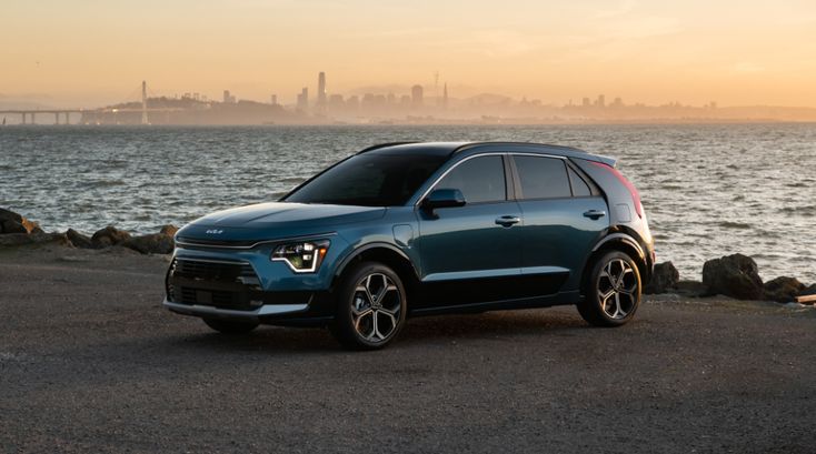 Blue SUV parked by the sea at sunset with city skyline in the background.