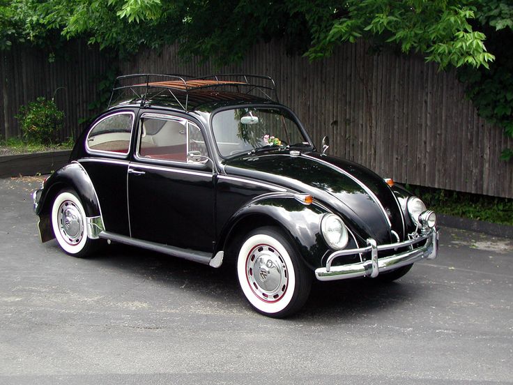 Classic black Volkswagen Beetle with roof rack parked on pavement, surrounded by greenery. Iconic vintage car design.