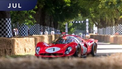 Red vintage racing car driving at speed on a tree-lined track during an event, flanked by hay bales and checkered flags.