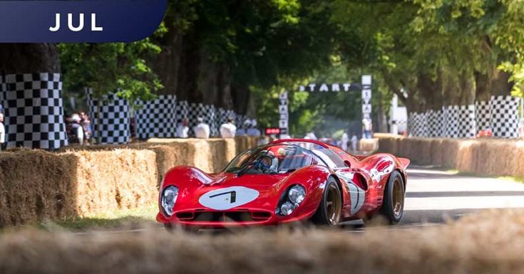 Red vintage racing car driving at speed on a tree-lined track during an event, flanked by hay bales and checkered flags.