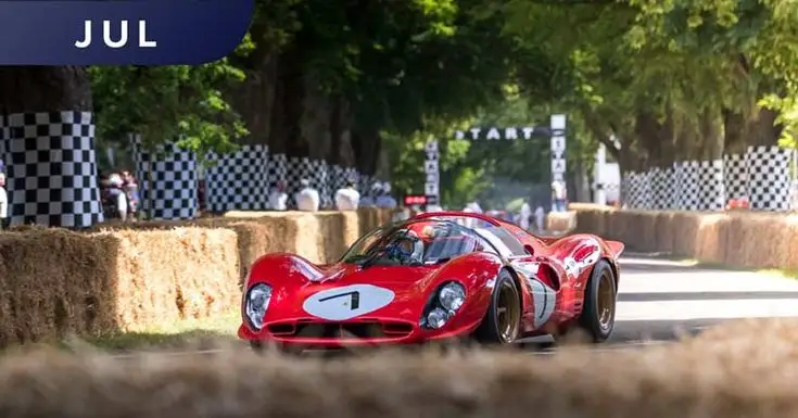Red vintage racing car driving at speed on a tree-lined track during an event, flanked by hay bales and checkered flags.