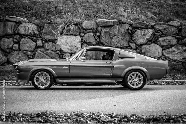 Classic vintage muscle car parked on a road beside a stone wall, black and white photograph.