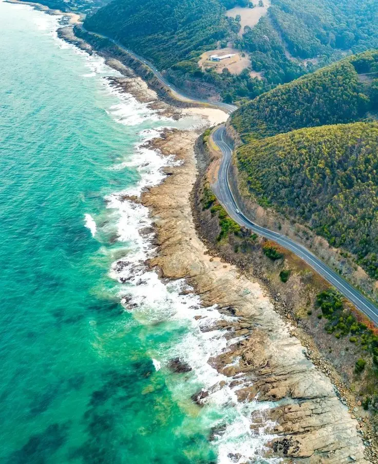 Aerial view of a coastal road winding along rugged cliffs and turquoise ocean waves, surrounded by lush green hills.