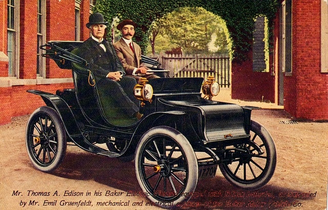 1910 Baker Electric Victoria with Thomas A_ Edison - Engine Stories Vintage car image showing two men seated in an early 20th-century electric vehicle, surrounded by brick buildings and greenery.