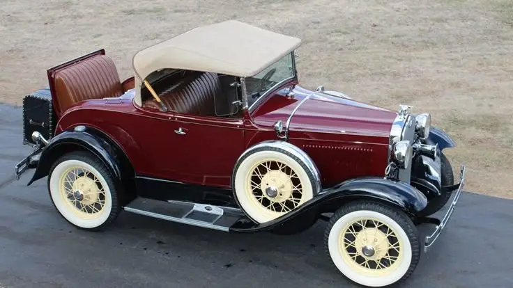 Vintage burgundy convertible classic car with a beige soft top, parked on a paved road with a grassy background.
