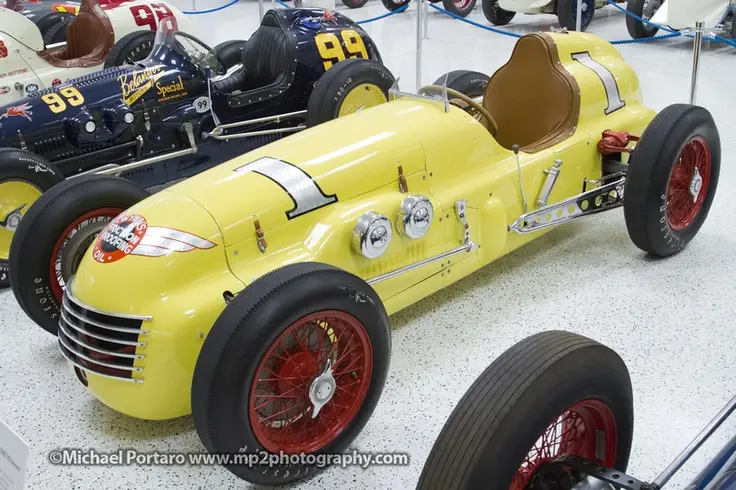 Yellow vintage race car with red wheels in a museum, surrounded by other classic racing vehicles, showcasing automotive history.