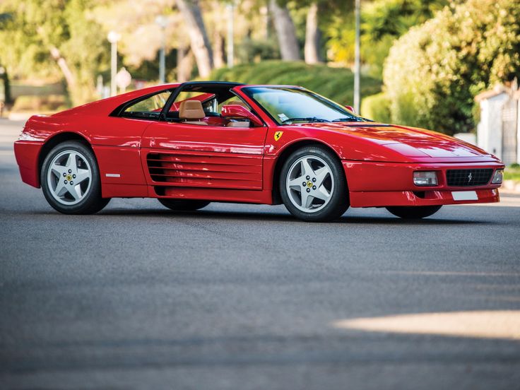 Red sports car parked on a road with lush greenery in the background. Classic car design with sleek curves and aerodynamic style.