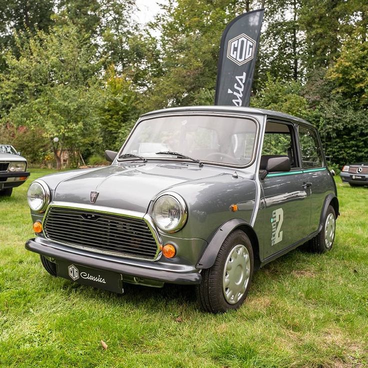 BMC Mini 1959 - Engine Stories Classic grey Mini Cooper on display at an outdoor event, surrounded by greenery and other vintage cars.