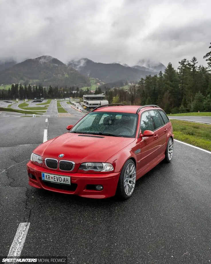 BMW E46 M3 Touring - Engine Stories Red BMW parked on a scenic mountain road with misty hills in the background. Ideal for car enthusiasts exploring nature routes.
