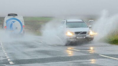 White SUV driving through flooded road, creating splash; van following behind in rainy weather.