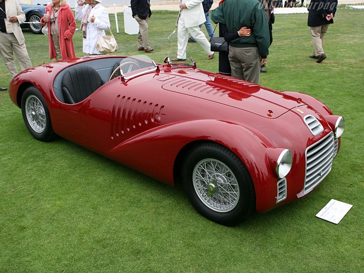 Red vintage sportscar displayed on grass at a classic car show, surrounded by spectators dressed in formal attire.