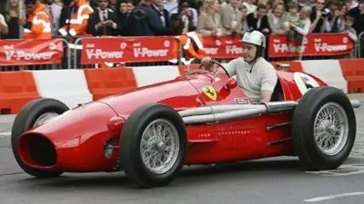 Vintage red Ferrari race car in action, driven by a helmeted racer on a street circuit. Crowd and barriers in the background.