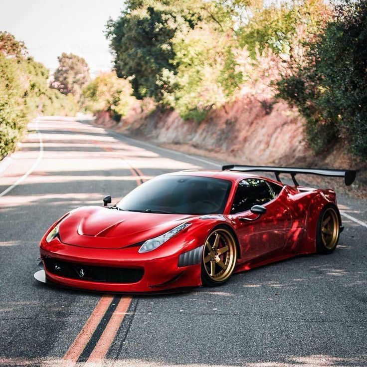 Red sports car with gold rims and large rear spoiler on a scenic, winding road surrounded by trees.