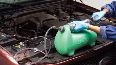 Mechanic using a coolant system refill kit on a car engine with the hood open.