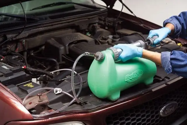 Mechanic using a coolant system refill kit on a car engine with the hood open.