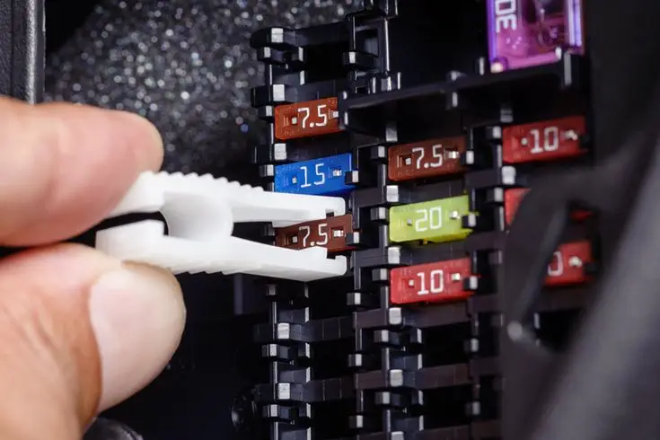 Hand using a tool to remove a fuse from a car's fuse box, showing various colorful fuses with different amperage ratings.