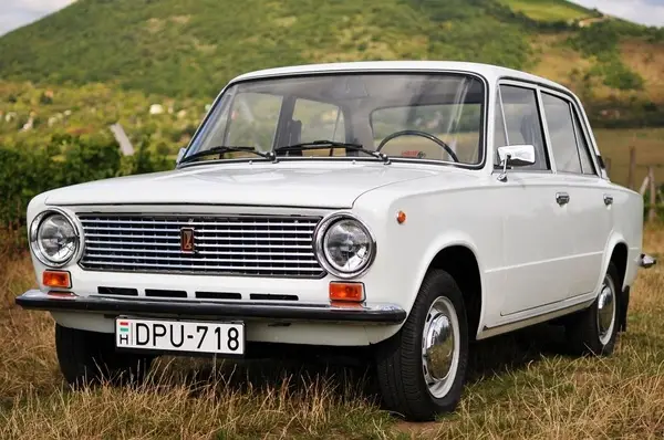 Lada 1970 - Engine Stories Classic vintage white car parked in a grassy field with green hills in the background.