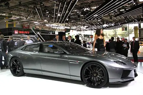 Lamborghini Estoque Concept - Engine Stories Sleek silver sports car showcased at an auto show with onlookers and ambient lights enhancing the vehicle's elegance and style.