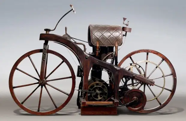 Maybach Daimler a wooden bicycle in 1885 - Engine Stories Antique wooden motorcycle, showcasing early engineering with large spoke wheels and a leather seat, against a neutral background.