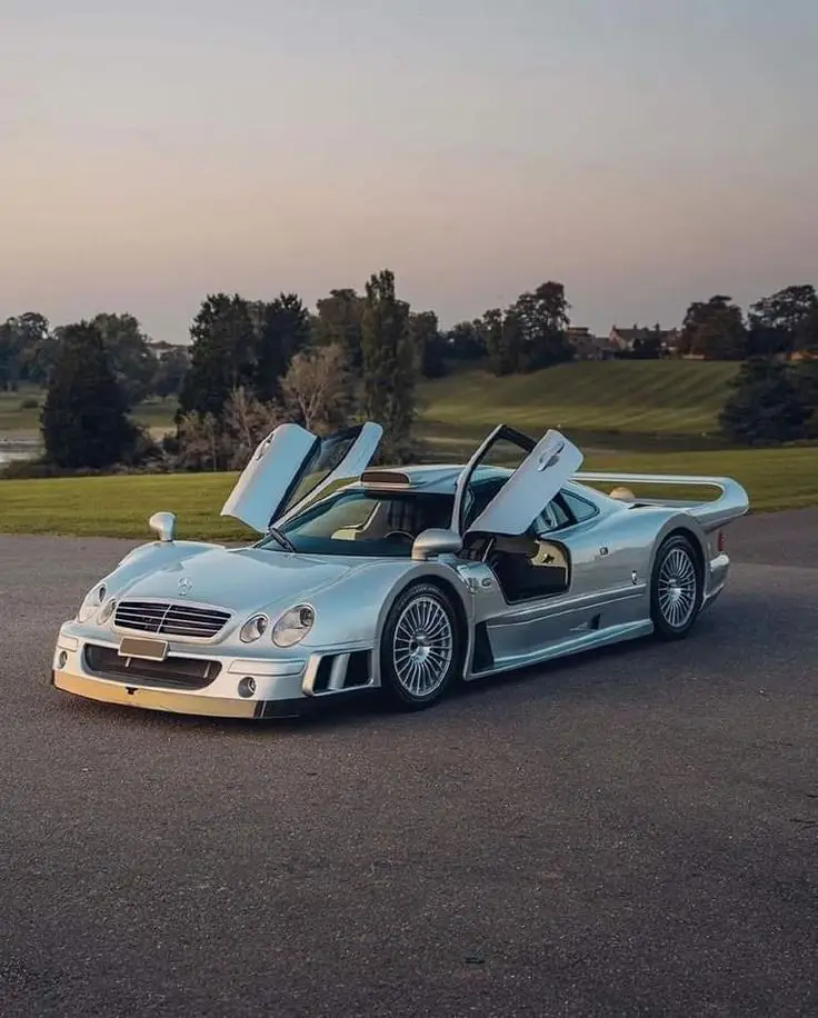 Mercedes CLK GTR - Engine Stories Silver luxury sports car with open gullwing doors, parked in picturesque outdoor setting at sunset.