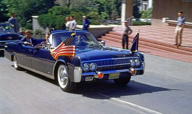 A classic blue convertible with flags drives down a city street, carrying passengers, surrounded by people and greenery in the background.