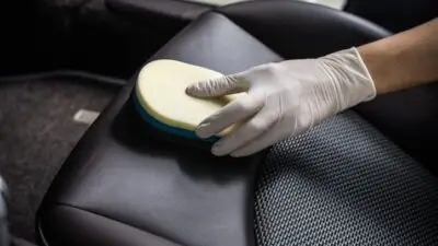 Gloved hand cleaning leather car seat with sponge for interior detailing and maintenance.