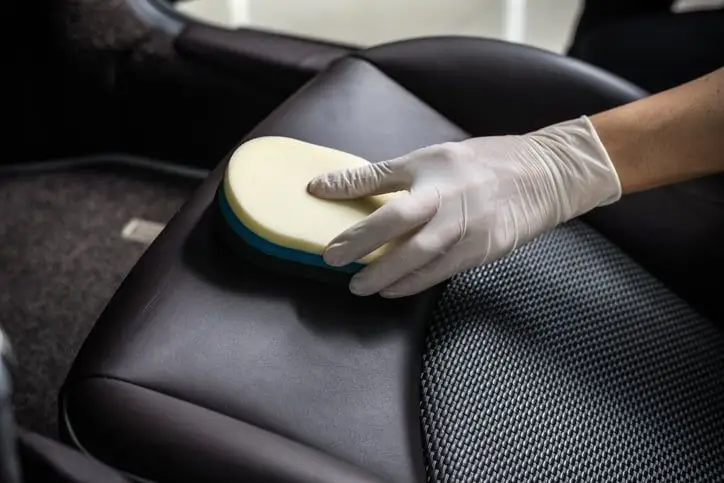 Gloved hand cleaning leather car seat with sponge for interior detailing and maintenance.