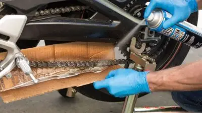 Person lubricating motorcycle chain with spray can, wearing blue gloves and using cardboard as a guard.