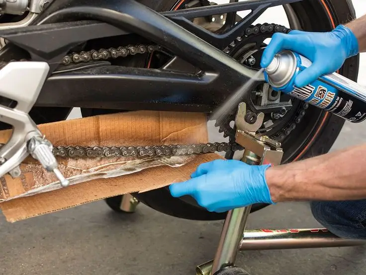 Person lubricating motorcycle chain with spray can, wearing blue gloves and using cardboard as a guard.