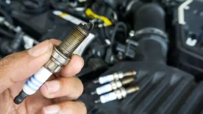 Hand holding a used car spark plug near engine, with three more plugs in the background. Ideal for automotive maintenance context.