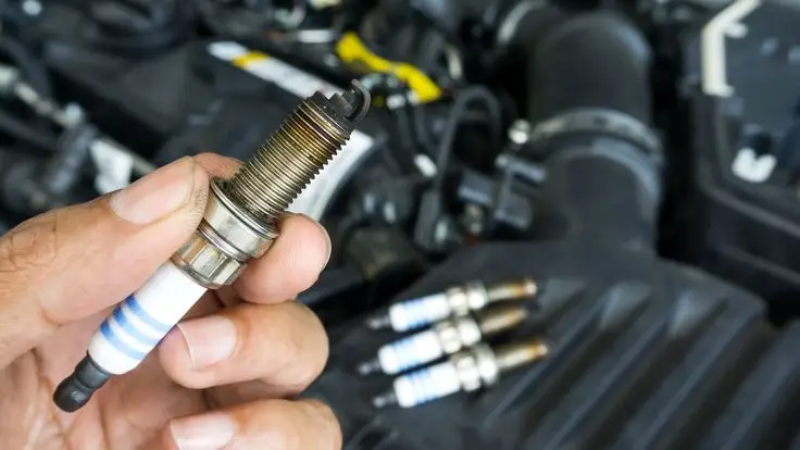 Hand holding a used car spark plug near engine, with three more plugs in the background. Ideal for automotive maintenance context.