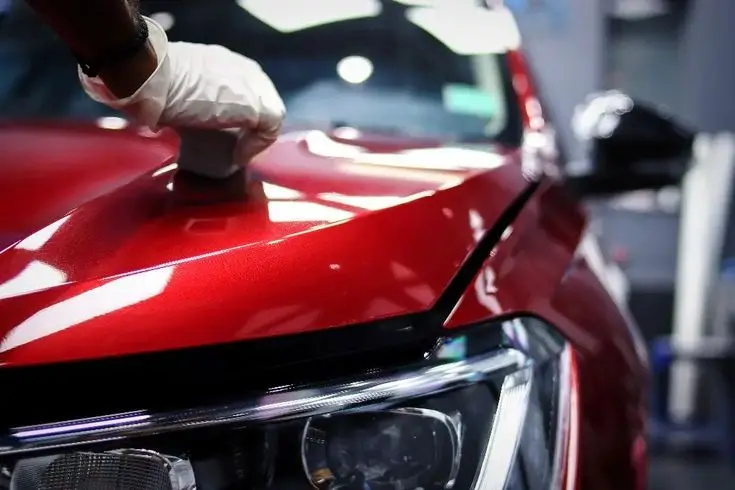 Gloved hand detailing a shiny red car hood in an auto workshop, emphasizing care and maintenance. Perfect for auto detailing content.
