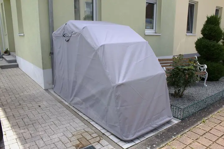 Car covered with a gray protective cover, parked beside a residential building on a paved area, near a small garden with a bench.