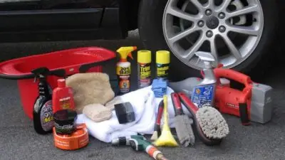 Car cleaning supplies including brushes, towels, and sprays arranged by a vehicle wheel on asphalt.